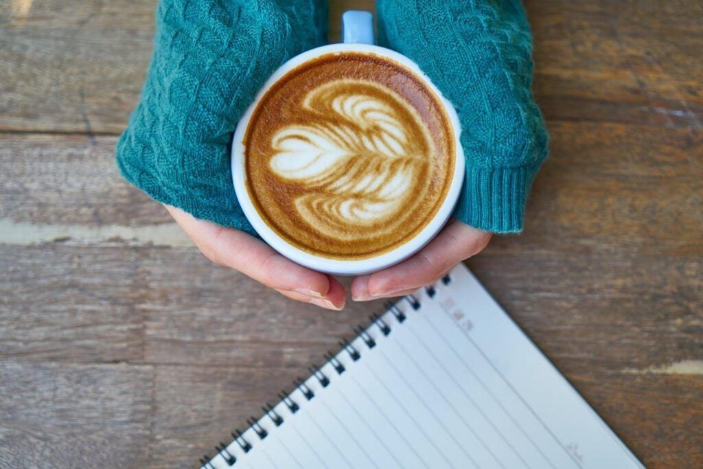 Alt.tag: a close-up of a woman’s hands holding a cup of coffee, next to an empty notebook